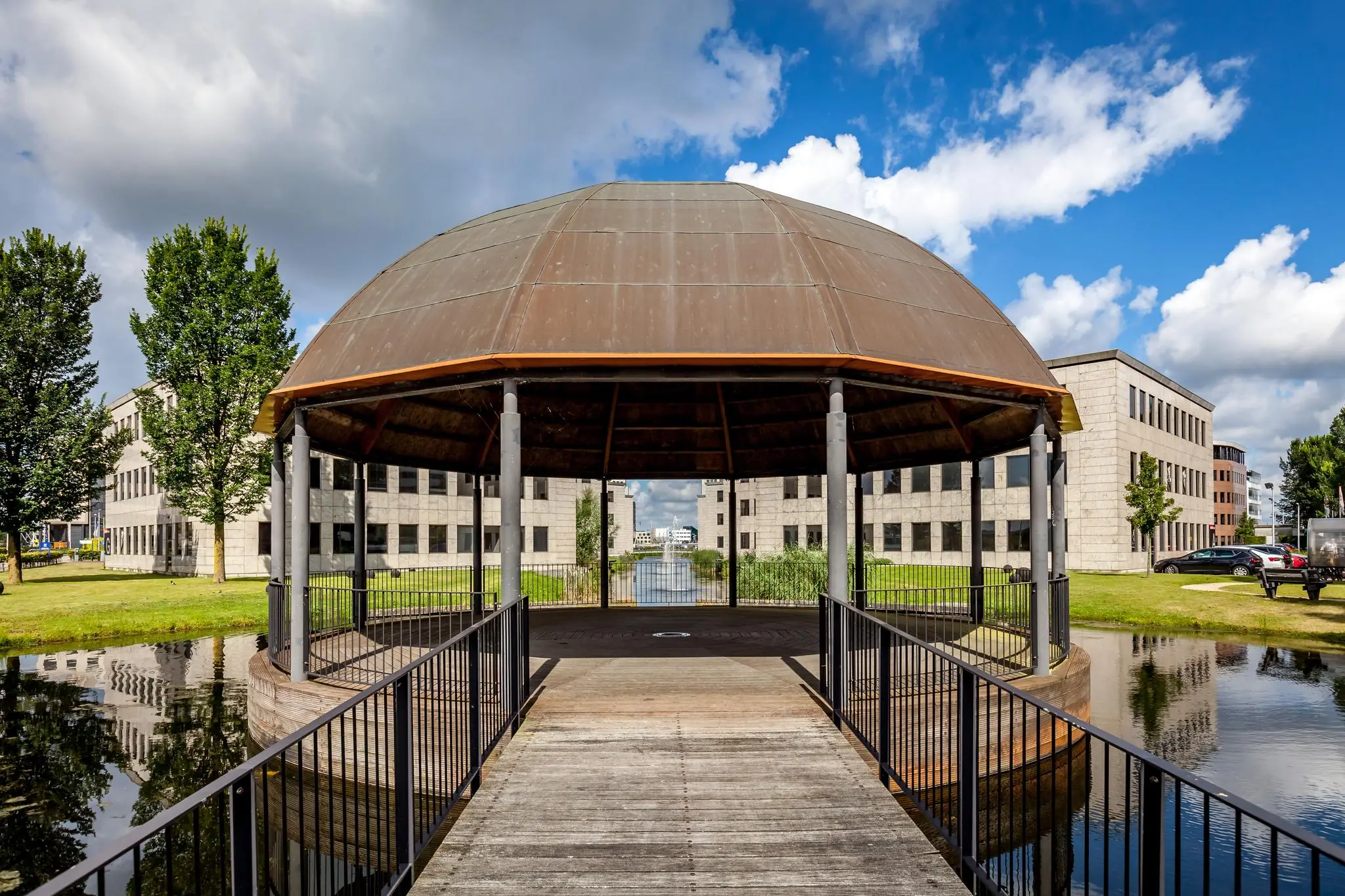 Een houten voetgangersbrug leidt naar een overdekt paviljoen aan de Rivium 1e Straat, omringd door moderne kantoorgebouwen en water.